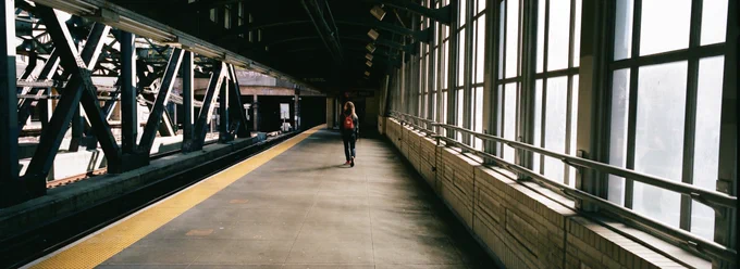 a sunlight elevated MTA train platform with a girl in the distance wearing a small red backpack and red chucks.