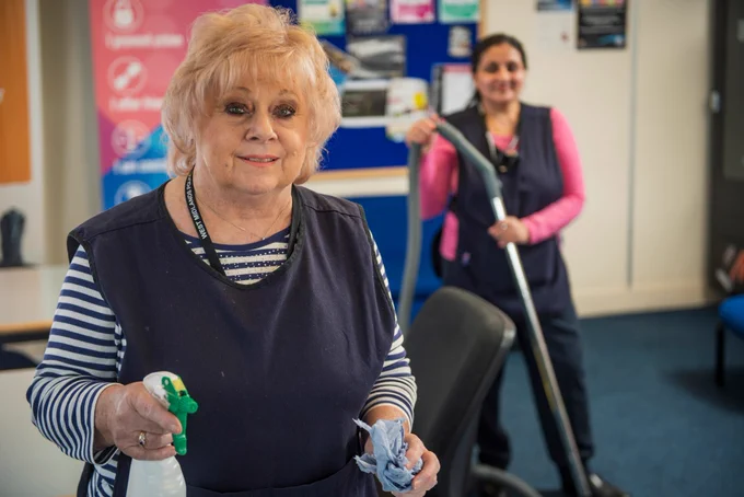 Two women are standing together. One is holding a hoover and the other is holding a spray bottle and cloth.