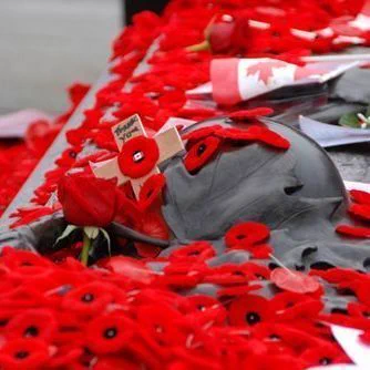 Color photograph of a war memorial covered in hundreds of red remembrance poppies. A black military helmet rests on the stone surface, surrounded by poppies, red roses, small Canadian flags, and handwritten notes. A wooden cross with a poppy is visible among the tributes. A person's hand appears on the left edge. The scene is outdoors on a gray stone memorial.