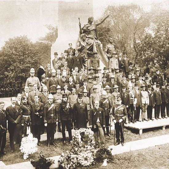 Sepia-toned photograph of a large group of men, many in military uniforms with medals and hats, posing in front of a tall stone war memorial topped with a statue of a winged figure holding a torch. The men stand in multiple rows on a platform and ground, some holding rifles. Floral wreaths are placed at the base. Trees and a crowd are visible in the background. The image shows age with scratches and fading.