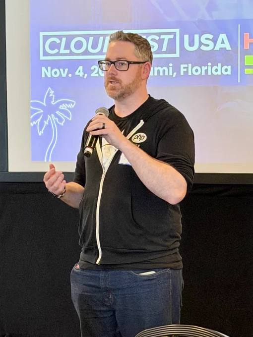 Scott K. Clark stands in front of a “CloudFest USA, Nov. 4, Miami, Florida” presentation screen, holding a microphone as they speak, wearing glasses, a black PHP hoodie, and jeans