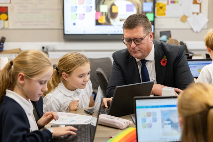 Sir Martyn Oliver in a classroom, talking to a female pupil using a laptop. 
