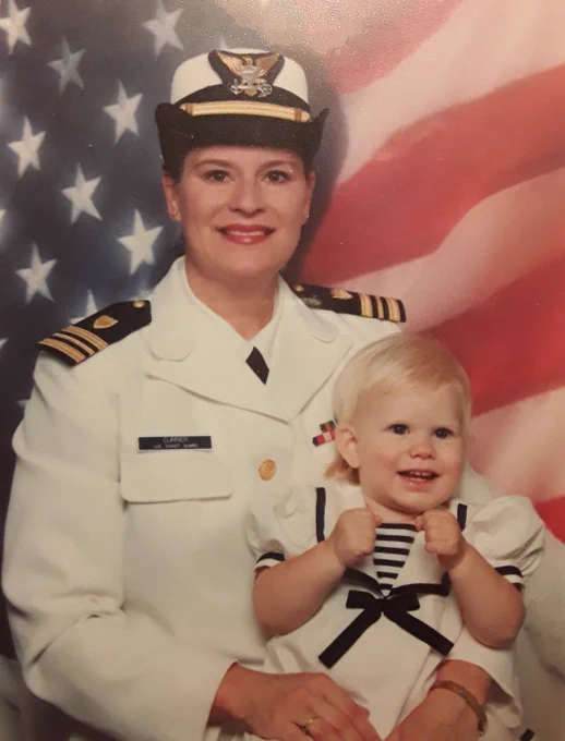 A person in U.S. Navy officer attire posing for a photo with a young child, both in front of an American flag. The child is wearing a sailor outfit.