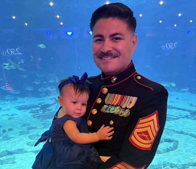 A person in a U.S. Marine Corps dress uniform holding a young child, standing in front of a large aquarium with various fish visible in the background.