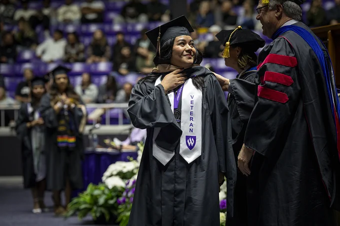A young brown woman graduates from Weber State University. She's wearing a white stole that says "Veteran" down the front in purple letters with the Weber State shield on it.