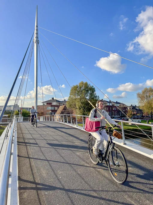 A woman in casual attire with a broom over her shoulder rides a bicycle across a car-free suspension bridge over a river in Rijswijk.