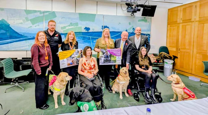 A group of ADI and ADUK members, along with Steve Darling MP, Lucy Edwards and Marc Abraham stand facing the camera with three assistance dogs at their feet