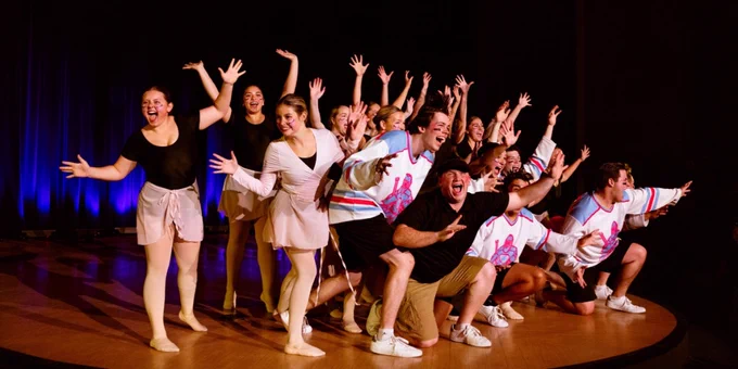 A group of enthusiastic performers in various costumes, including cheerleading outfits and dance attire, enthusiastically posing on stage under theatrical lighting, conveying energy and excitement in their performance.