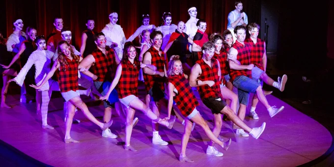 A group of dancers wearing red and black plaid shirts and white pants performs a dynamic ensemble dance routine on stage under dramatic lighting.