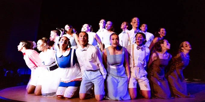 Cast of performers on stage in a theatrical production, dressed in various pastel and white costumes, looking upwards with expressive faces, under dramatic lighting.