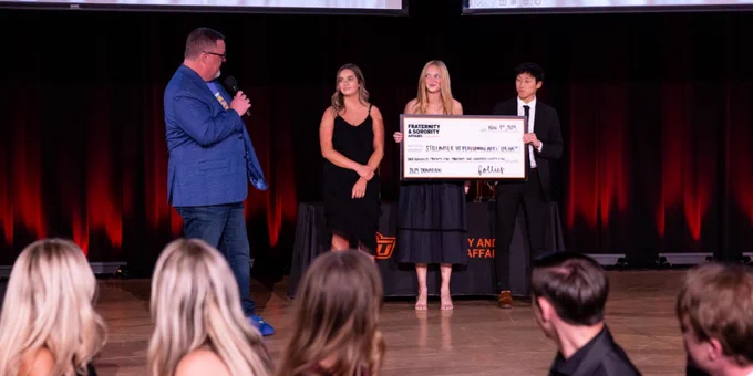 Three individuals, one holding a large ceremonial check from Oklahoma State University, stand onstage as another person speaks into a microphone during an event.