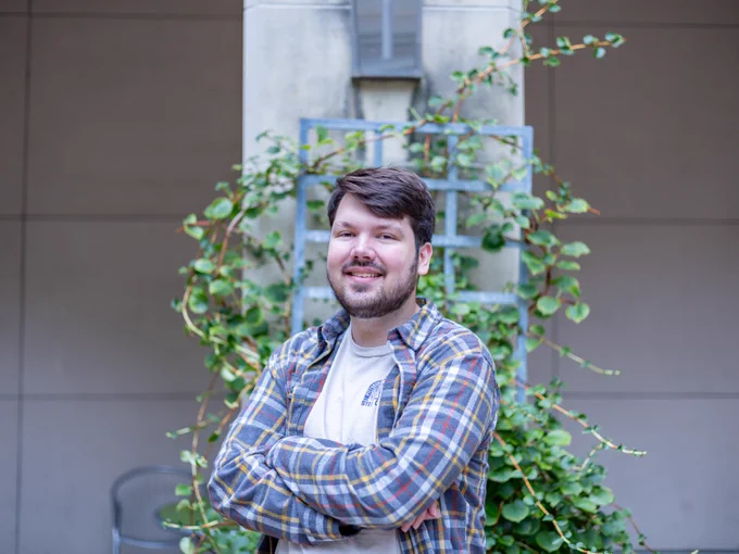 A person wearing a plaid shirt standing with crossed arms and smiling in front of a vine-covered wall.