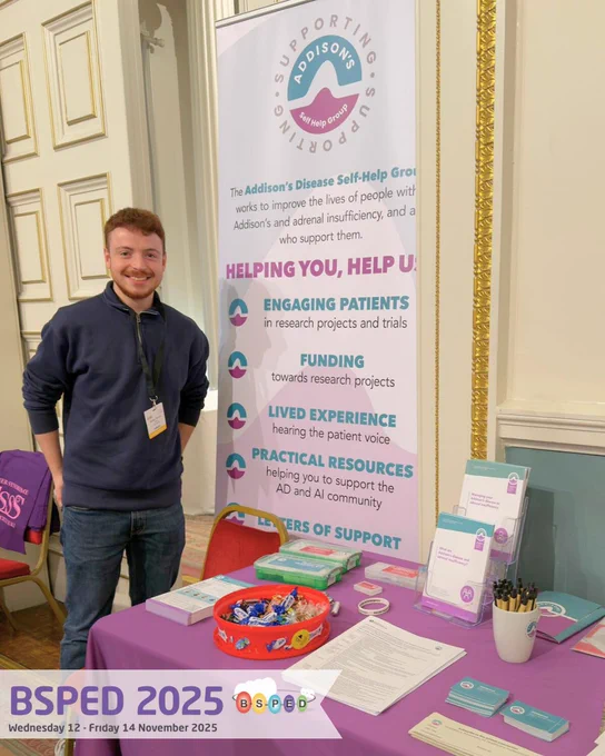 A smiling man in a blue jumper and jeans stands beside an Addison’s Disease Self-Help Group (ADSHG) display table at the BSPED 2025 conference. 

The stand features a large banner outlining the charity’s aims, with the subheader "Helping You, Help Us". Bullet points list engaging patients, funding, lived experience, practical resources, and letters of support. 

The table is covered with an ADSHG branded purple cloth and displays leaflets, information packs, pens, and a bowl of chocolates!

The BSPED 2025 logo and event dates (Wednesday 12 – Friday 14 November 2025) are visible in the bottom corner.