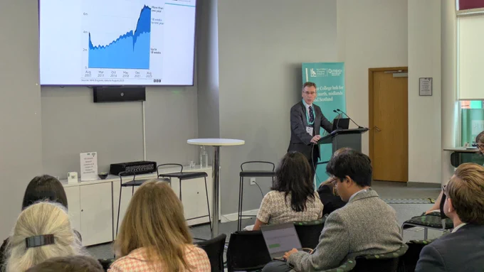 RCS England President, Tim Mitchell, delivers an address to an audience. There is a screen with a chart behind him.