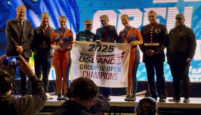 A group of eight people on stage holding a "2025 USBANDS GROUP IV OPEN CHAMPIONS" banner.