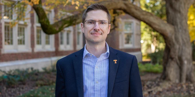 Christopher Stripling wearing glasses, a plaid shirt, and a blazer with a University of Tennessee pin, standing in front of a brick building with autumn trees.