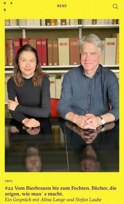 A woman (on the right) and a man (on the left) sit at a table, facing forward; the background is shelving with archives and rare books.