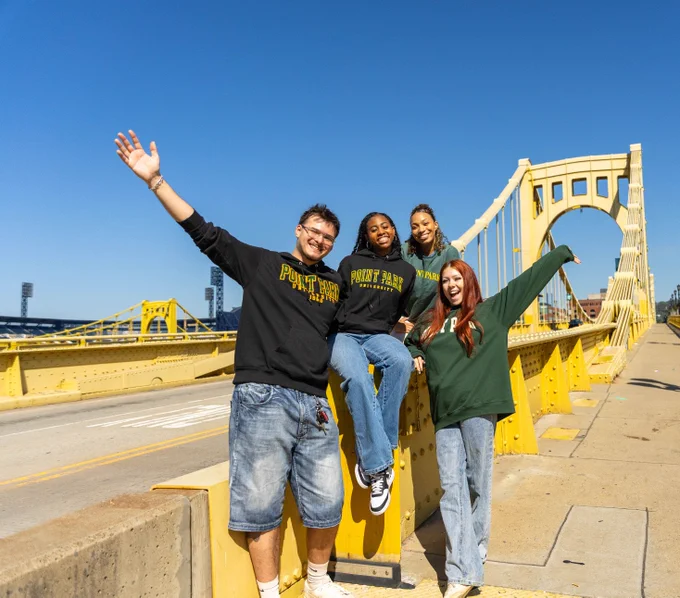 Four students wearing Point Park University sweatshirts joyfully posing on one of the three sister bridges.