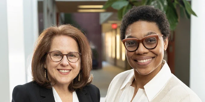 Two individuals smiling in a corporate hallway, one with shoulder-length hair wearing glasses and a black blazer, and the other with short curly hair wearing glasses and a beige blazer.