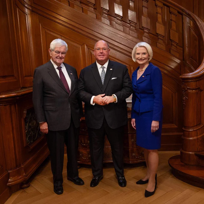 Ambassador Callista L. Gingrich and Speaker Newt Gingrich pose for a photo with former Ambassador Edward McMullen from the American Swiss Foundation in front of a wooden staircase.