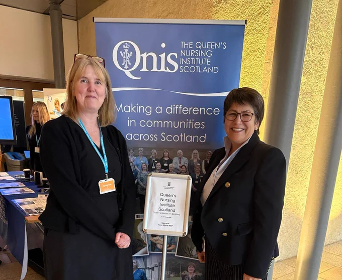 Two people standing indoors in front of a display for The Queen’s Nursing Institute Scotland. The display banner reads, ‘Making a difference in communities across Scotland.’ Both individuals are smiling and wearing professional attire with name badges. A small sign beside them also references the Queen’s Nursing Institute Scotland.