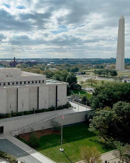 Aerial view of the National Museum of American History with the Washington Monument in the background, surrounded by greenery under a partly cloudy sky.