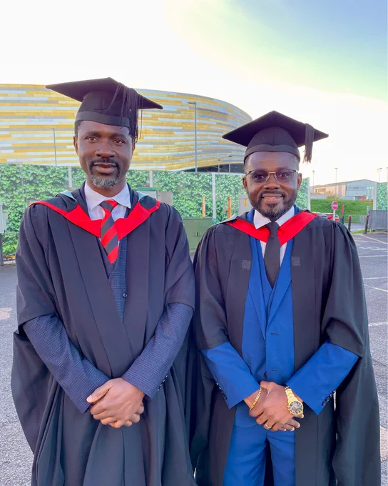 Two graduates in black gowns and mortarboards stand side by side outdoors with a Derby Arena in the background.