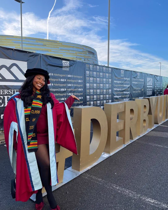 Graduate in maroon gown and colorful stole posing beside large gold 'DerbyUni' letters outdoors under blue sky.