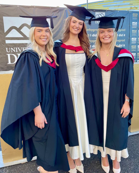 Three female graduates wearing black gowns and mortarboards stand together in front of a university banner outdoors.