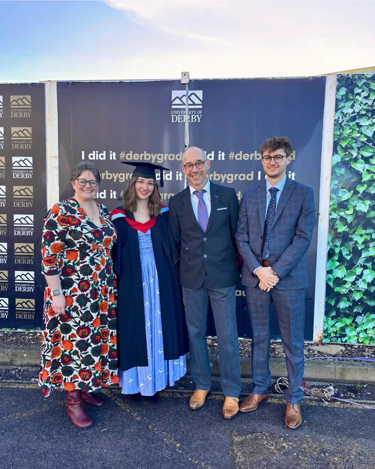 Four people posing in front of a University of Derby graduation backdrop, one wearing a graduation gown and cap.