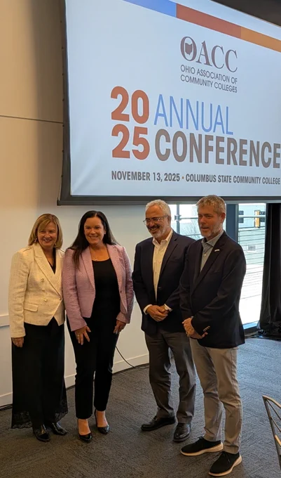 The college president and other panelists (another man and two women in business attire) smile. 