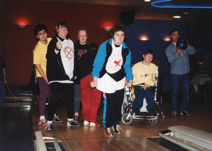 Six young people, some with disability conditions, standing or sitting, all playing or watching ten pin bowling at indoor bowling alley.
