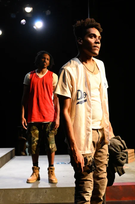 Two actors on stage in a dramatic scene, with one standing in the foreground looking slightly off to the side and another watching from the background. Both are dressed in casual, modern attire under stage lighting.