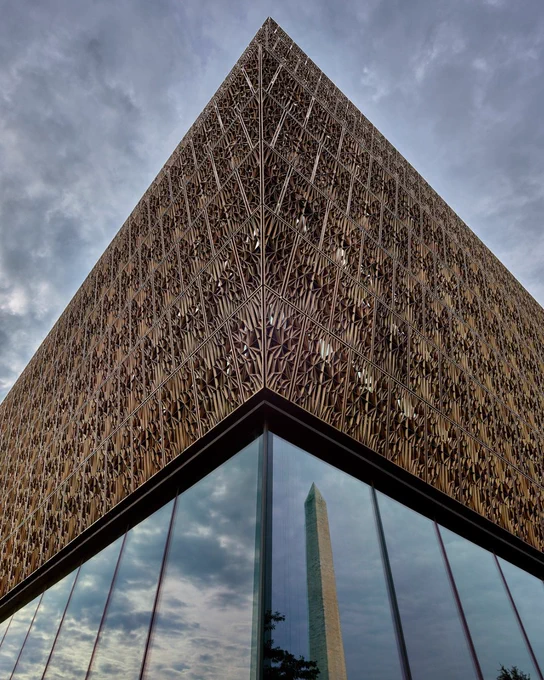 Low angle view of the intricate lattice facade of the National Museum of African American History and Culture with the Washington Monument reflected in the glass.