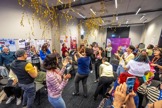 
This is a celebratory photograph from the Bi  Equal Conference and Founding Meeting.

A large group of people are gathered indoors in a conference room, cheering, clapping, and taking photos as gold metallic streamers fall from the ceiling. Many people are smiling and laughing, creating a feeling of excitement and success. The attendees are diverse in age and appearance. Some attendees are holding or wearing items in the colors of the bisexual and pansexual pride flags. Posters and banners, including one reading "Bi  Equal Founding Meeting," are visible on the surrounding walls.