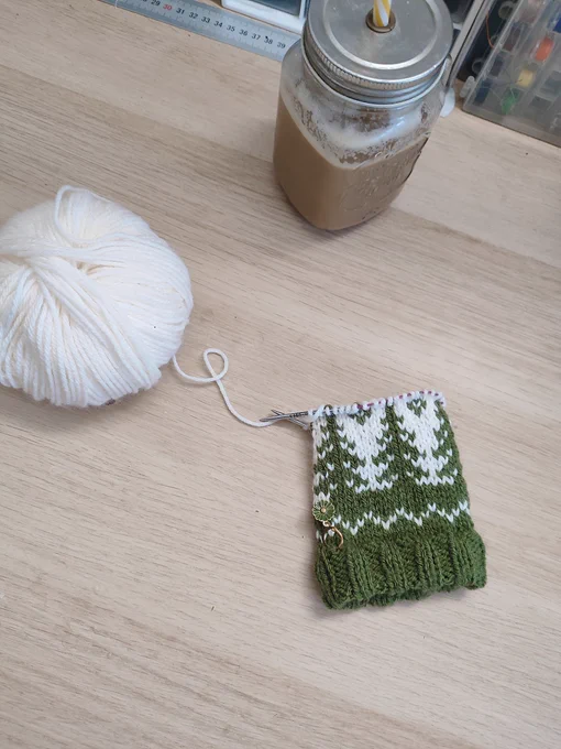 Photo de mon bureau avec le début d'un tricot de ce même motif, encore attaché à la pelote de fil blanc et avec mon mug de café en haut 