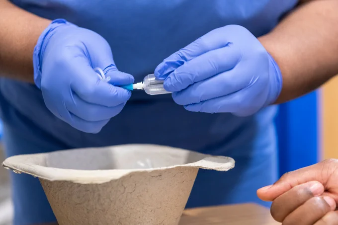 A person wearing blue gloves is holding a syringe above a bowl, preparing to inject or dispense a substance, with another hand visible nearby.