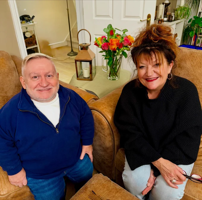 Bright pic with Jane on the right in a black soft jumper and smiling. Simon smiling too with white tee underneath a navy quarter zip top. Both on sofas flowers in the middle.