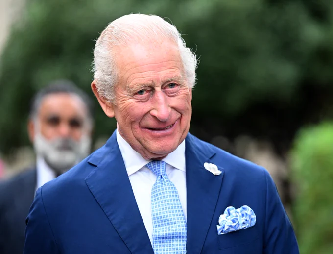 His Majesty The King wearing a blue suit and blue tie and smiling into the camera - trees can be seen in the background.