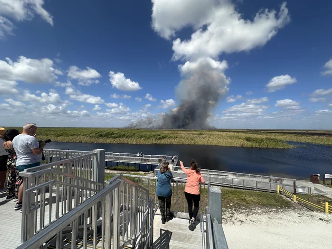 People on a boardwalk watching thick black smoke rising from a fire across a river in a grassy wetland area under a partly cloudy sky.