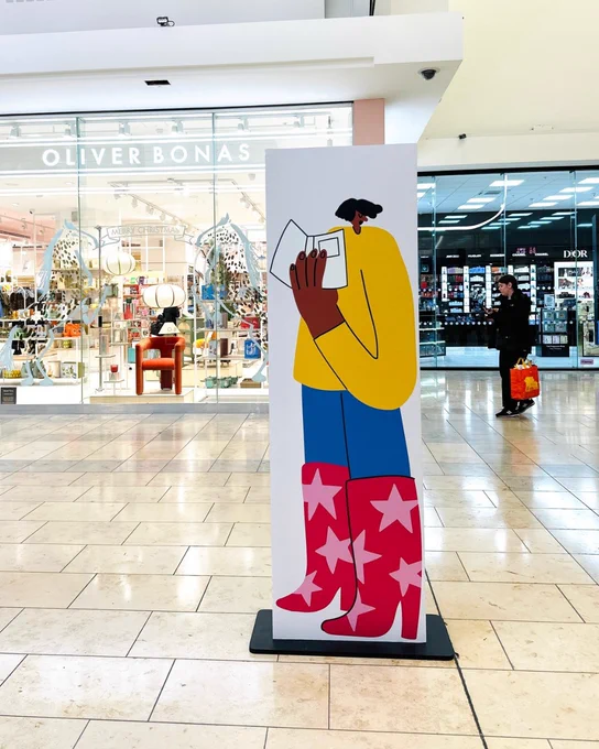 A standee in a shopping mall featuring an illustrated figure reading a book, with a large Oliver Bonas store in the background.