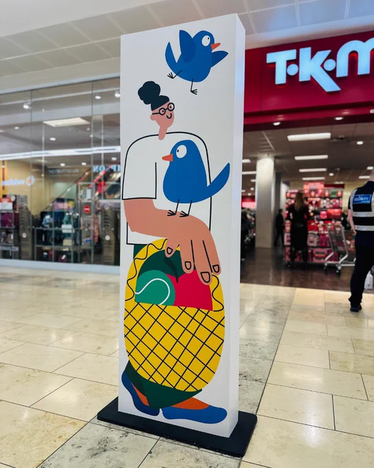 Promotional stand featuring a playful illustration of a person holding a pineapple with a large blue bird perched on their hand, located in a shopping center with a TK Maxx store in the background.
