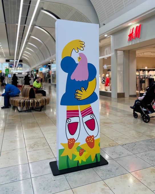 A colourful digital kiosk displaying a cartoon graphic of a whimsical character with a blue body and yellow head, positioned in a lively shopping centre near the H&M store.