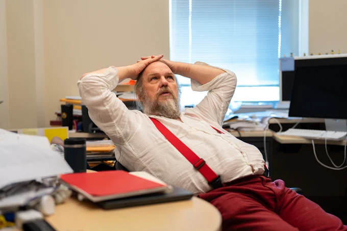 A bearded man in a white shirt and red suspenders leans back in a cluttered office, appearing thoughtful or stressed.
