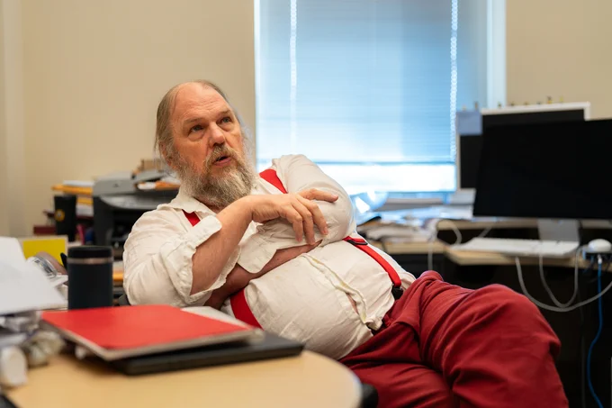 A bearded man with red suspenders sits in an office, gesturing while speaking. A computer and office supplies are visible in the background.