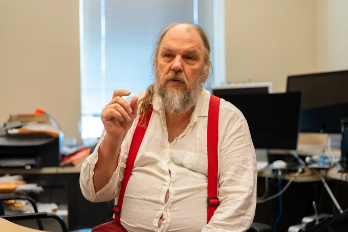 An older man with a long white beard and hair, wearing a white shirt and red suspenders, gestures while seated in an office with computers.
