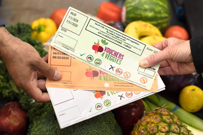 Close-up of two people exchanging colorful “Vouchers 4 Veggies” paper coupons over a table of fresh produce, including broccoli, peppers, bananas, and a pineapple.