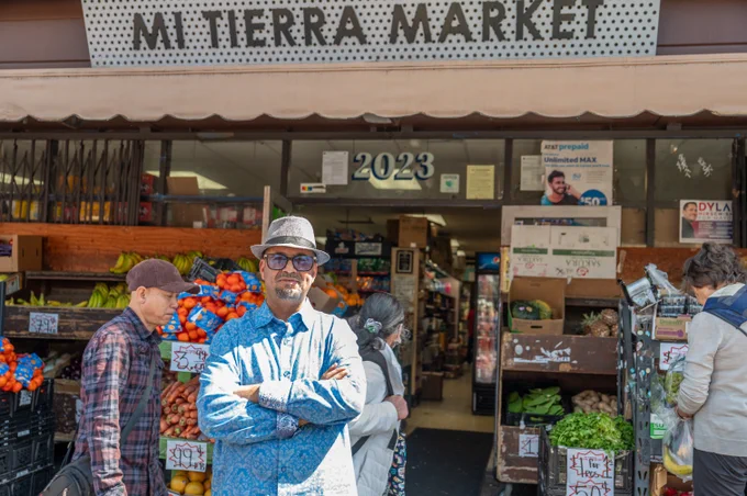 A man wearing a blue shirt, sunglasses, and a gray fedora stands with his arms crossed outside Mi Tierra Market. Fresh fruits and vegetables are displayed around him as shoppers walk by the storefront.