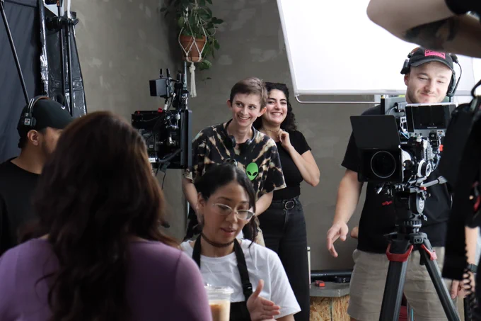 Many crew members in the background running the camera and monitor, smiling at cast members seated in the foreground. 
