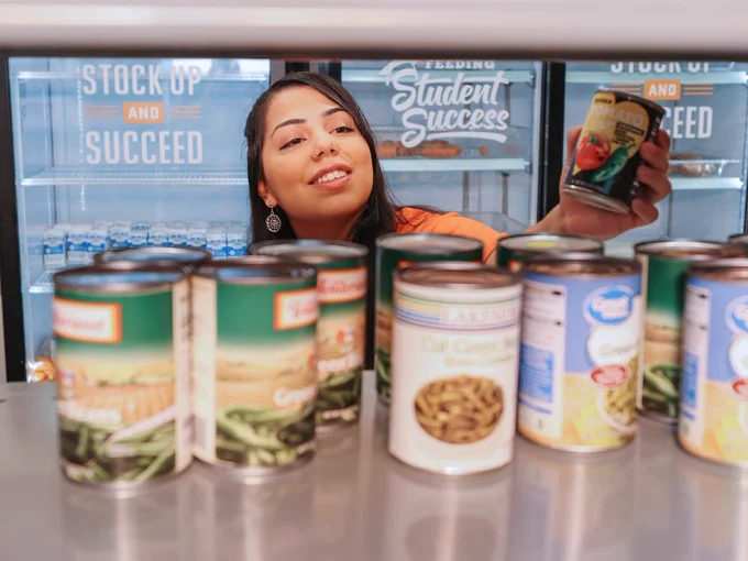 Person smiling while stocking a shelf with assorted canned goods in a Student Food Pantry.